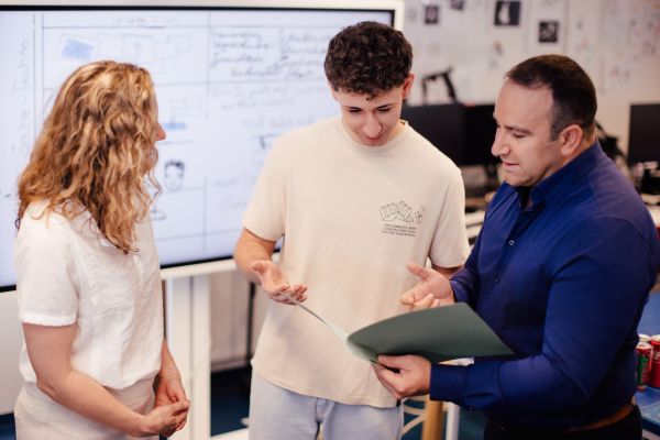 Berufsausbildungsassistentin, ein Berufsschüler und ein Berufsschullehrer stehen vor einer digitalen Tafel; der Lehrer zeigt ein Dokument her.